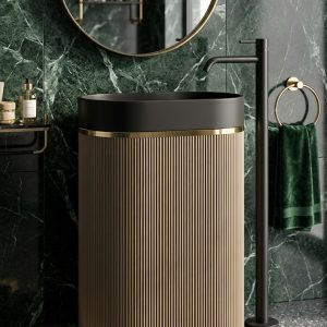 Muniq artificial stone pedestal basin 1341 600x375x850mm showing warm sand fluted oval column pedestal with matte black oval bowl and precision gold ring against dark green wall with planting
