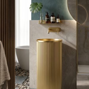 Flute Gold freestanding stainless steel pedestal basin with brushed gold PVD fluted cylinder form glowing warm gold in dark bathroom with chevron tiles and round LED mirror