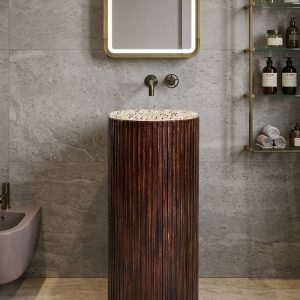 Muniq artificial stone pedestal basin with deep walnut wood effect fluted cylinder pedestal and white terrazzo bowl rim in warm biophilic bathroom with travertine walls and brass mirror